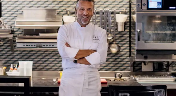 Chef Perbellini, sorridente in cucina, con le braccia conserte. Dietro di lui, attrezzature professionali e un forno acceso.