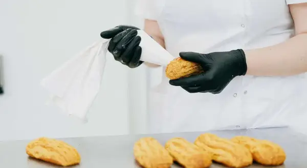 Ręce w czarnych rękawiczkach przygotowują ptysie do nadzienia kremem z żółtek. Na stole leżą gotowe ciastka.