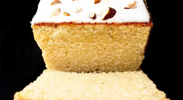 A slice of almond cake with white icing and chopped almonds on top, against a black background.
