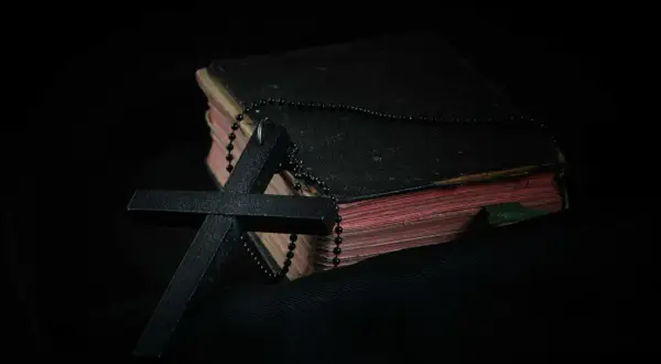 Czarny krzyż i paciorki różańca na starej księdze. Symbol wiary i modlitwa o spokój ducha w mroku.