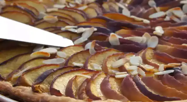 Une tranche de tarte aux nectarines, garnie d'amandes effilées, est coupée par un couteau.