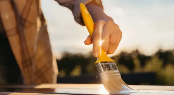 Mano che stende vernici poliuretaniche bicomponenti su un tavolo di legno al tramonto, per una finitura protettiva e duratura.