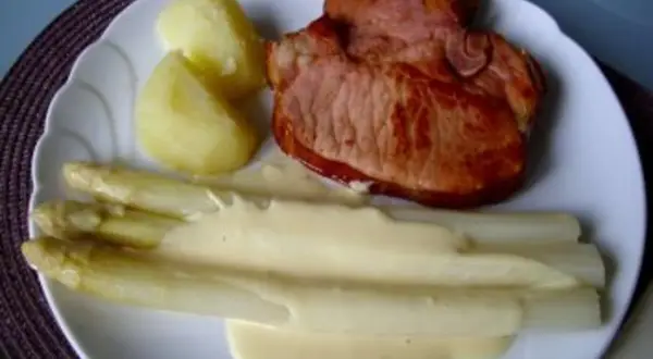 Ein Teller mit gebratenem Schinken, Kartoffeln und Spargel in Sauce Hollandaise. Der Spargel ist in Alufolie gewickelt.