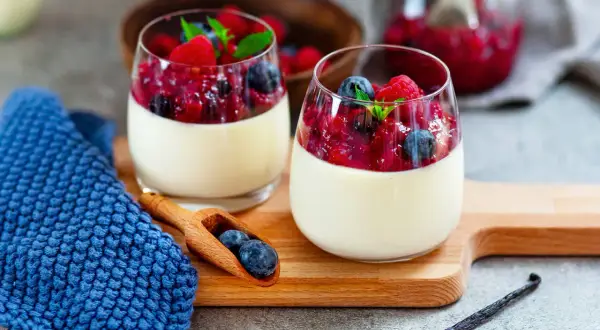 Zwei Gläser cremiger Panna cotta mit Beerenkompott und frischen Beeren, garniert mit Minze. Ein Löffel mit Blaubeeren liegt daneben.