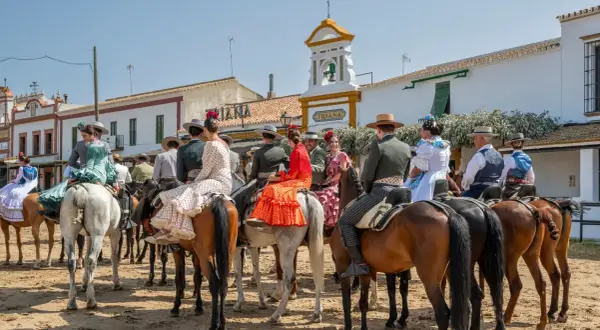 Gente vestida de flamenco a caballo, en un **camino rocío** bajo un cielo azul. Edificios blancos y un campanario al fondo.