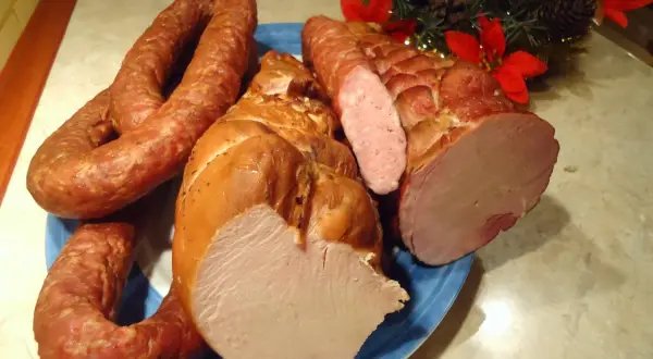 Wędzona szynka i kiełbasa na niebieskim talerzu, ozdobione gałązką choinki.