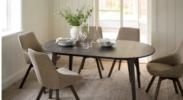 Ett runt mörkt matbord med fyra stoppade stolar runt sig. En vas med blommor, ljusstakar och en skål med frukt på bordet. En tidlös design klassiker.