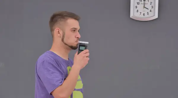 Młody mężczyzna dmucha w alkomat, sprawdzając, ile promili ma po lampce szampana. Zegar na ścianie pokazuje czas.
