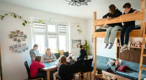 Rodzina gra w planszówkę w domu pomocy społecznej "Przystań". Dzieci siedzą na łóżku piętrowym, obserwując grę.