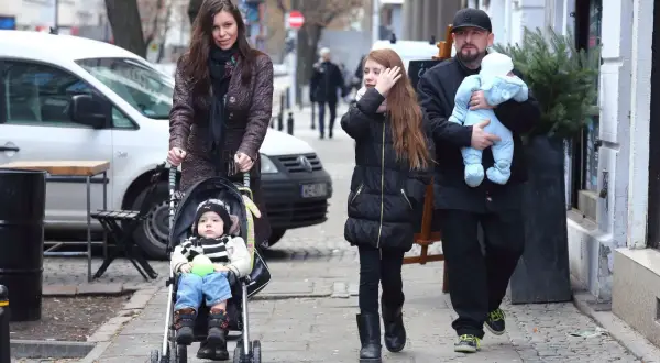Rodzina spaceruje po mieście, mama pcha wózek z niemowlakiem, a tata niesie drugie dziecko. Wszyscy liroy dzieci wyglądają na szczęśliwych.