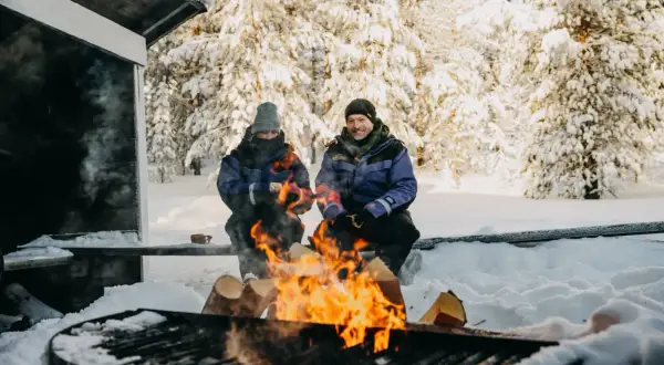 Un couple profite d'un feu de camp dans un paysage de Laponie hivernale, entouré d'arbres enneigés.