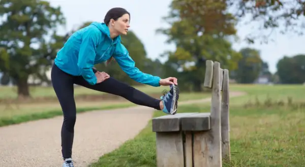Donna in abbigliamento sportivo blu esegue un riscaldamento pre corsa, allungando la gamba su una panchina in un parco.