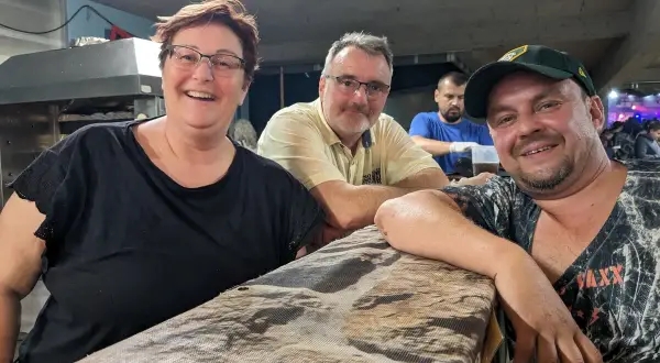 Fröhliche Menschen am holzmarkt ilsfeld. Eine Frau mit Brille und schwarzem Shirt, ein Mann mit gelbem Hemd und ein Mann mit Kappe lächeln in die Kamera.