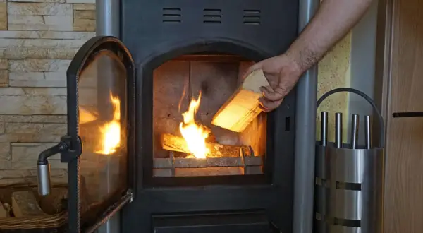 Hand legt Holzscheit mit gutem Brennwert Birke in den brennenden Ofen.