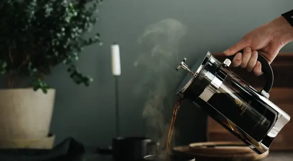 Här hälls nybryggt kaffe till kaffepressen i en mörk mugg. Ångan stiger från den varma drycken.