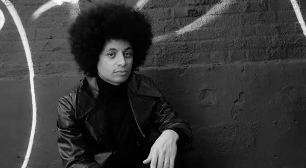 Jose James z burzą afro, w skórzanej kurtce i golfie, siedzi na tle ściany z graffiti.