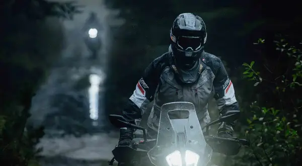 Motocyklista w kasku i kurtce jedzie po mokrej, leśnej drodze w nocy.