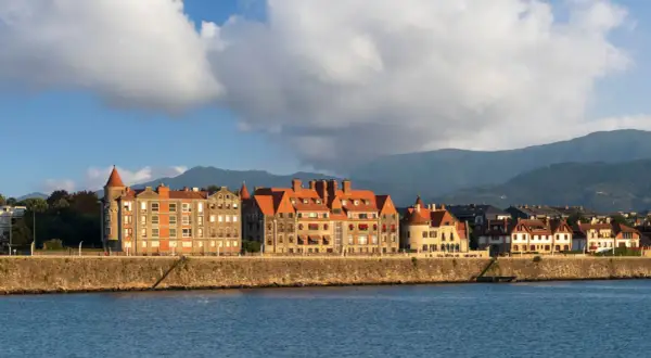 Edificios históricos con tejados rojos bordean el paseo de las grandes villas Getxo, con montañas al fondo y el mar en primer plano.