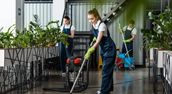 Dziewczyna w granatowym stroju do sprzątania odkurza podłogę. W tle inni pracownicy dbają o czystość i zieleń.