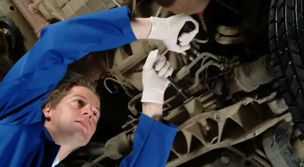 Mechanik w niebieskim kombinezonie naprawia samochód, używając klucza do dokręcenia śruby. Jego biała manszeta jest widoczna.