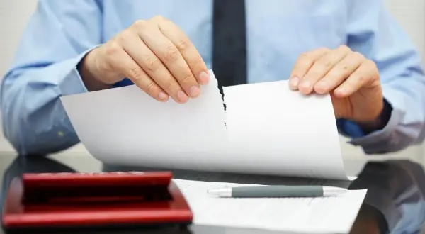 Uomo in camicia blu strappa un documento, simbolo di annullamento proposta di acquisto immobile. Calcolatrice e penna sul tavolo.