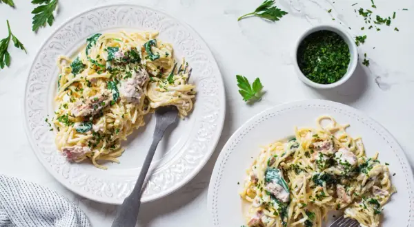 Dwa talerze spaghetti z kremowym sosem, szpinakiem i kawałkami tuńczyka z puszki, posypane natką pietruszki.