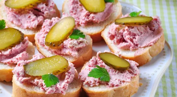 Kromki chleba z pyszną pastą z szynki, ozdobione plasterkami ogórka kiszonego i natką pietruszki. Idealna przekąska!