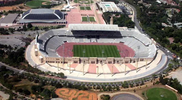 Widok z lotu ptaka na stadion olimpijski Montjuïc z zieloną murawą i trybunami. W pobliżu inne obiekty sportowe i tereny zielone.