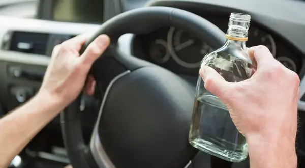 Ręce kierowcy na kierownicy, jedna trzyma butelkę alkoholu. To niebezpieczne zachowanie, które łamie limit alkoholu w Polsce.