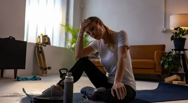 Erschöpfte Frau sitzt auf einer Matte, Hand an der Stirn. Vielleicht ein Fehler beim Muskelaufbau? Trainingsgeräte liegen herum.
