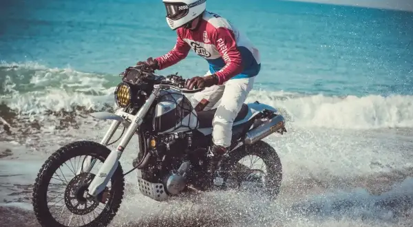 Motocyklista w kasku i kolorowej bluzie śmiga na Honda FMX 650 po plaży, wzbijając fontanny wody.