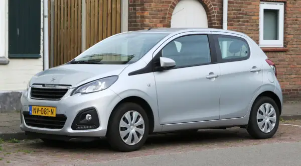 Srebrny Peugeot 108 stoi zaparkowany na brukowanym podjeździe, obok ceglanego domu.