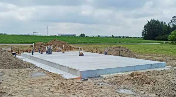 Gotowa płyta fundamentowa na budowie, otoczona piaskiem i zielenią. W tle widać budynki przemysłowe.