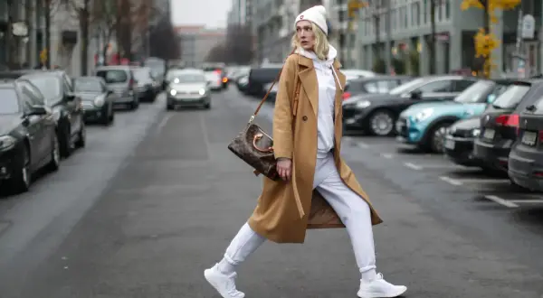 Kobieta w beżowym płaszczu i białych dresach oversize przechodzi przez ulicę, niosąc torebkę.
