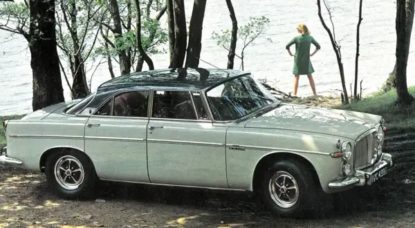 En ljusgrå Rover P5 står parkerad vid en sjö. En kvinna i grön klänning står vid vattenbrynet.