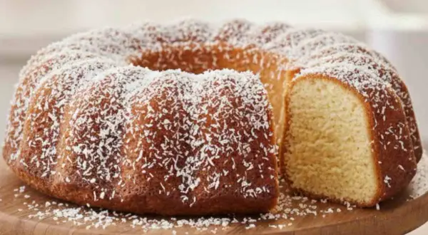 Ciambella al cocco dorata e soffice, cosparsa di scaglie bianche su un tagliere di legno.