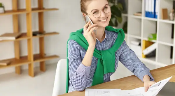 Uśmiechnięta kobieta w okularach prowadzi rozmowę telefoniczną, przeglądając dokumenty. To może być część jej instruktażu stanowiskowego.
