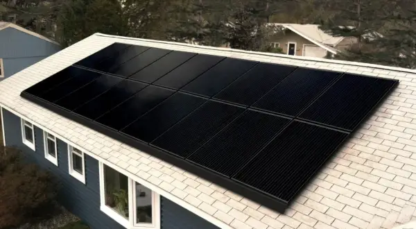 Paneles negros de una aplicacion placas solares cubren el tejado de una casa azul, rodeada de árboles.