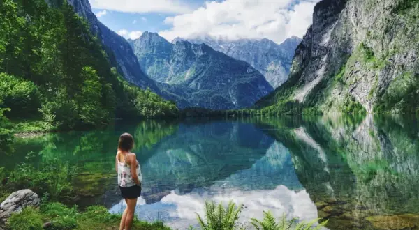 Frau blickt auf spiegelglatten See in den bayerischen Regionen, umgeben von steilen, bewaldeten Bergen.
