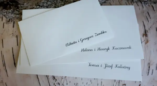 Białe koperty ślubne z elegancko wypisanymi nazwiskami gości na tle kory drzewa.