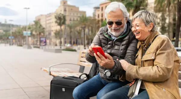 Uśmiechnięta para seniorów na ławce w mieście przegląda telefon. Czyżby sprawdzali, co to roaming, planując kolejną podróż?