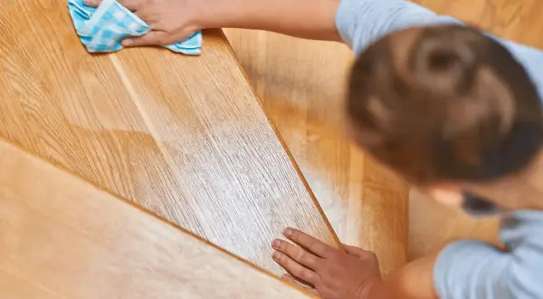 Une main nettoie un escalier en bois verni encrassé avec un chiffon bleu à carreaux.