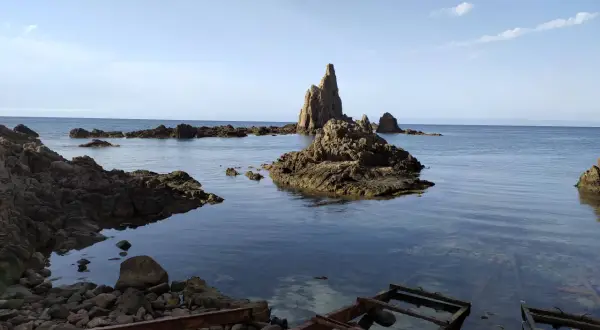 Rocas emergiendo del mar azul, como el arrecife de las sirenas. El agua cristalina revela el lecho rocoso y restos oxidados en la orilla.