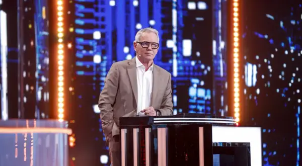 Mężczyzna w garniturze stoi przy pulpicie na tle sceny z niebieskimi światłami.
