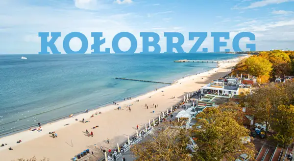 Widok na plażę w Kołobrzegu z ludźmi, budynkami i morzem.