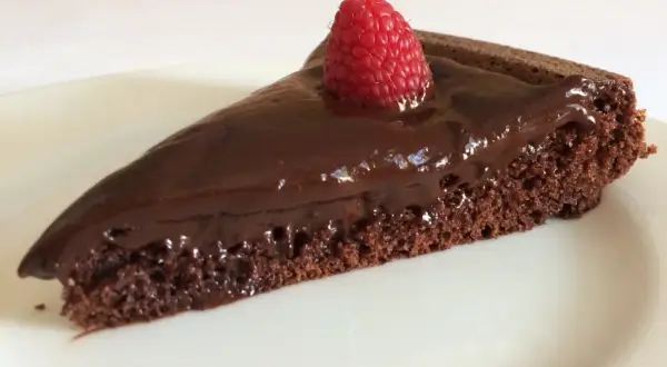 Una fetta di gateau al cioccolato, lucida e golosa, con una fragola fresca in cima, su un piatto bianco.