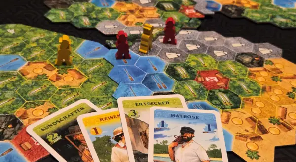 Spielbrett mit Figuren und Karten. Die Karten "KUNDSCHAFTER", "REISEN", "ENTDECKER" und "MATROSE" sind ausgelegt. Eine deutsche Spielepreis Liste könnte diese Elemente zeigen.