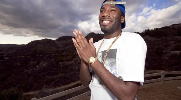 Dougie F. smiles and claps his hands, wearing a blue cap and gold chains, with a dramatic cloudy sky and hills behind him.