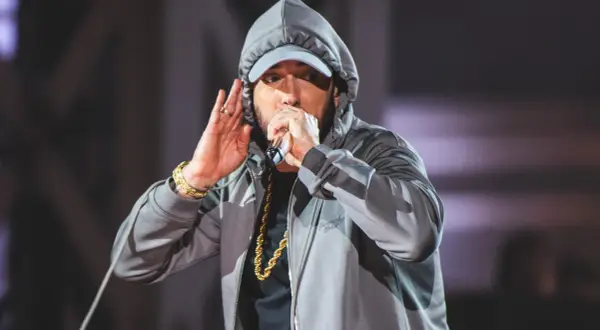 Eminem w szarej bluzie z kapturem i czapce, trzymający mikrofon i gestykulujący.
