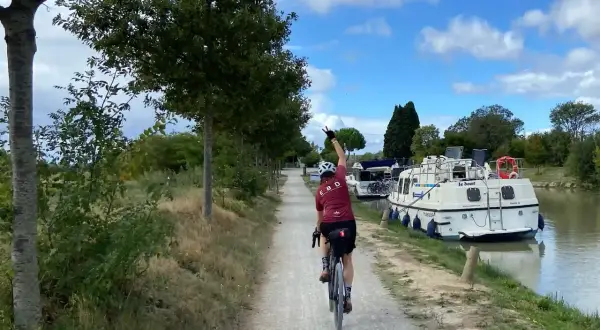 Ciclista pedaleando por el Canal du Midi, con barcos amarrados a un lado y árboles al otro.